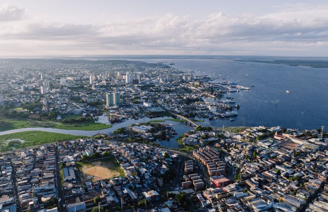 Bird's-eye view of São Raimundo cityscape, featuring river, buildings, and harbor under a cloudy sky.